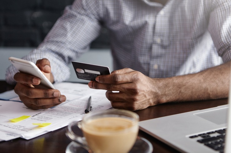 Un uomo seduto a un tavolo controlla il saldo del conto corrente sul cellulare tenendo la carta di credito in mano