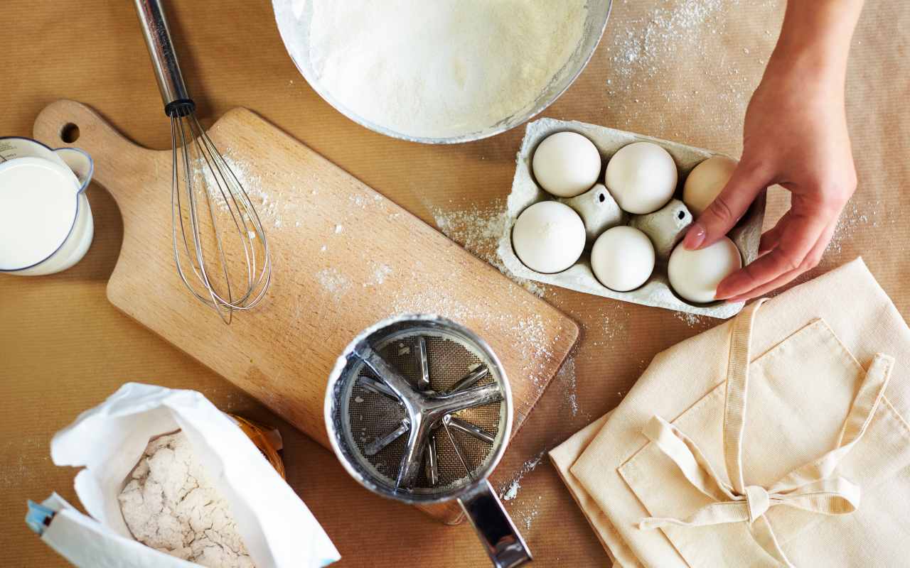 Una persona prepara un dolce con uova, latte, farina e utensili da cucina sul tavolo